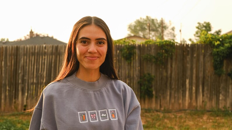 May include: A person wearing a gray sweatshirt with four small, square embroidered designs. The designs include a carrot, a bow, a book, and a bottle. The background features a wooden fence and green foliage.