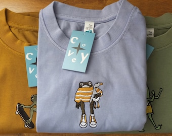 May include: Three t-shirts with different embroidered designs. The center shirt is light blue with a frog wearing a striped shirt and holding a goose. The left shirt is mustard yellow with a frog riding a skateboard. The right shirt is olive green with a frog wearing shorts.