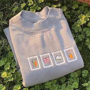 May include: A gray sweatshirt with a ribbed crew neck and embroidered postage stamp designs. The stamps feature a book, a bow, an open book, and a vase of flowers. The sweatshirt is folded on a bed of green foliage.