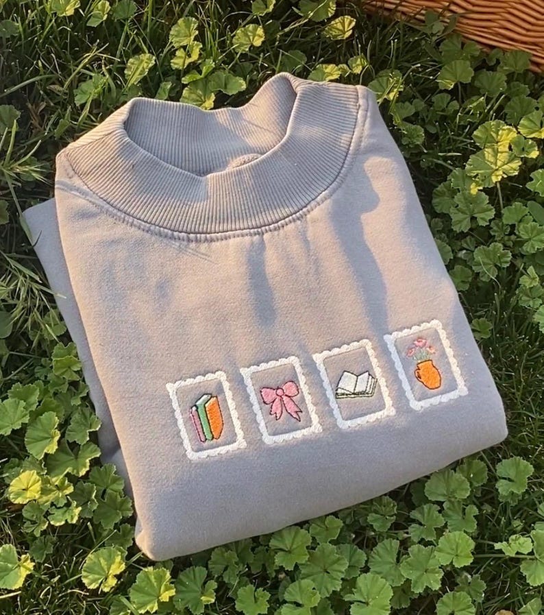 May include: A gray sweatshirt with a ribbed crew neck and embroidered postage stamp designs. The stamps feature a book, a bow, an open book, and a vase of flowers. The sweatshirt is folded on a bed of green foliage.