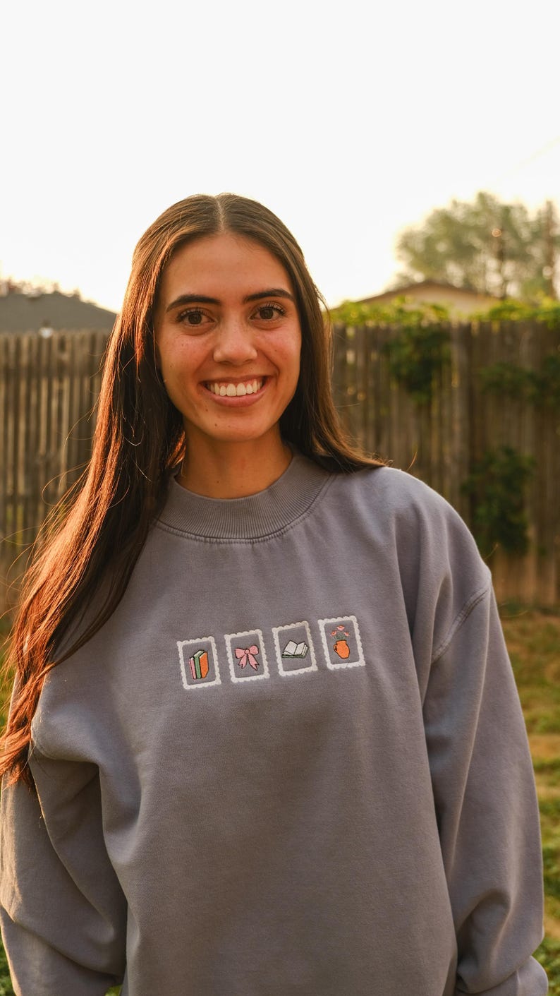 May include: A gray sweatshirt with embroidered postage stamp designs. The stamps feature a carrot, a pink bow, a book, and a peach. The sweatshirt is worn by a person with long brown hair, smiling in front of a wooden fence.