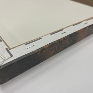 May include: Close-up of a stretched canvas art print. The canvas is stapled to a wooden frame with a brown and tan patterned edge. The canvas is white and the staples are silver.