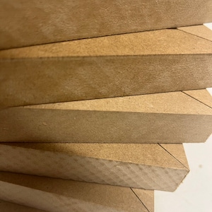 May include: Close-up of a stack of unfinished wooden boards. The boards are a light brown color and have a rough, textured surface. Each board has a beveled edge, creating a V-shape at the corner. The boards are stacked neatly.