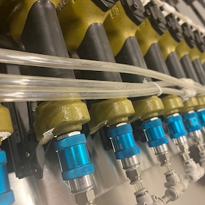 May include: Close-up of a series of industrial spray nozzles. The nozzles have a metallic olive green finish with black rubber tips. Blue and silver quick-connect fittings are visible, along with clear plastic tubing. The equipment is mounted on a metal surface.