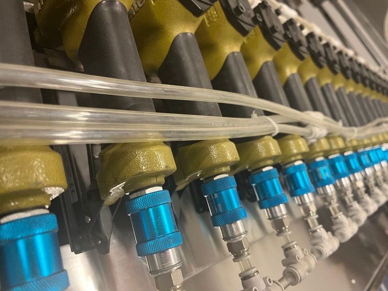 May include: Close-up of a series of industrial spray nozzles. The nozzles have a metallic olive green finish with black rubber tips. Blue and silver quick-connect fittings are visible, along with clear plastic tubing. The equipment is mounted on a metal surface.