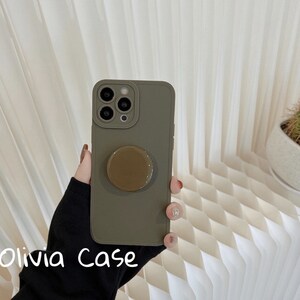 May include: A green phone case with a pop socket attached. The case is made of a soft, flexible material and has a matte finish. The pop socket is brown and has a smooth, glossy finish.