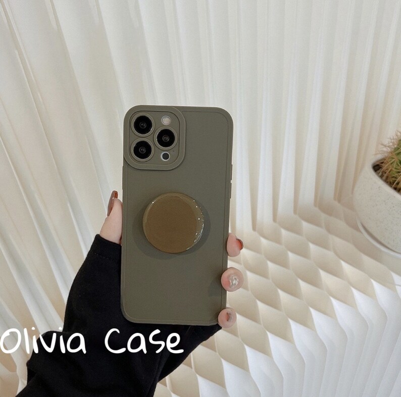 May include: A green phone case with a pop socket attached. The case is made of a soft, flexible material and has a matte finish. The pop socket is brown and has a smooth, glossy finish.
