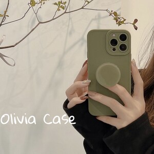 May include: A sage green phone case with a pop socket attached. The case has a camera lens cutout and the text "Olivia Case" is written on the bottom of the image.