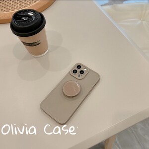 May include: A beige phone case with a pop socket attached. The case has a camera cutout with three lenses. The text "Olivia Case" is written on the table below the phone case.