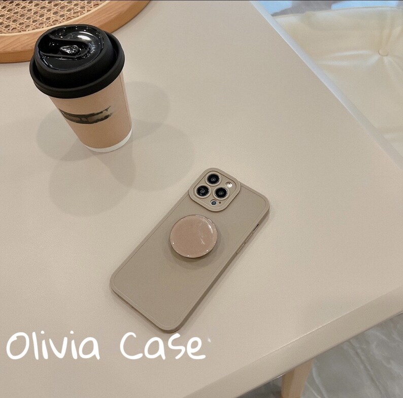May include: A beige phone case with a pop socket attached. The case has a camera cutout with three lenses. The text "Olivia Case" is written on the table below the phone case.