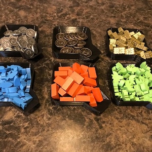 May include: Six black trays filled with game pieces. The trays contain silver coins, gold coins, wooden blocks, blue fish-shaped pieces, orange rectangular blocks, and green leaf-shaped pieces. The trays are on a brown surface.