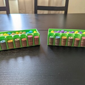 Puede incluir: Dos organizadores de pl&aacute;stico verde con compartimentos para guardar peque&ntilde;as piezas de juego. Cada organizador tiene una fila de huevos de pl&aacute;stico de colores y una pila de piezas de juego marrones, amarillas y verdes.