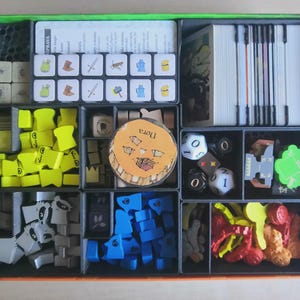 May include: A board game box organiser containing various game components including stacks of white cards, a grid of small square tiles with icons, yellow and blue wooden shapes, grey raccoon-shaped tokens, dice, and a circular token labelled Nora.