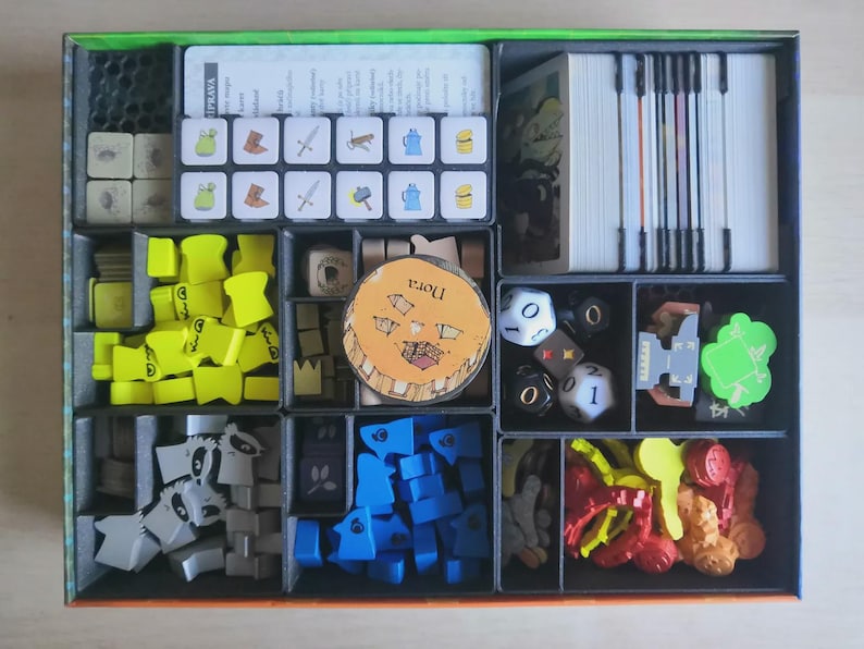 May include: A board game box organiser containing various game components including stacks of white cards, a grid of small square tiles with icons, yellow and blue wooden shapes, grey raccoon-shaped tokens, dice, and a circular token labelled Nora.