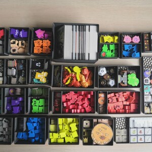 May include: A board game organiser set featuring multiple black plastic trays filled with various colourful wooden game pieces, tokens, dice, and card decks. The components are organised by colour and shape, including red, blue, green, yellow, and purple items.