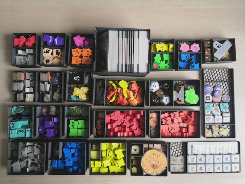 May include: A board game organiser set featuring multiple black plastic trays filled with various colourful wooden game pieces, tokens, dice, and card decks. The components are organised by colour and shape, including red, blue, green, yellow, and purple items.
