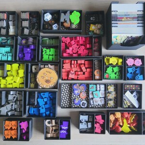 May include: A board game organiser set consisting of multiple dark grey plastic trays filled with various game components, including colourful wooden tokens, small cards, dice, and circular markers, all neatly arranged in separate compartments.
