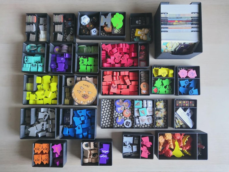 May include: A board game organiser set consisting of multiple dark grey plastic trays filled with various game components, including colourful wooden tokens, small cards, dice, and circular markers, all neatly arranged in separate compartments.