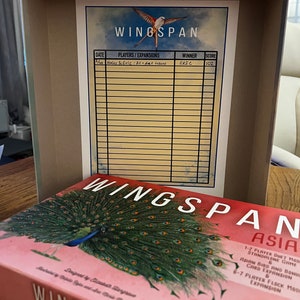 Puede incluir: Una caja de juego de mesa Wingspan con un fondo rosa y una ilustraci&oacute;n de un pavo real. La caja tiene el texto "Wingspan Asia" y un logotipo de Stonemaier Games. La caja est&aacute; abierta y se puede ver una hoja de puntuaci&oacute;n en el interior.