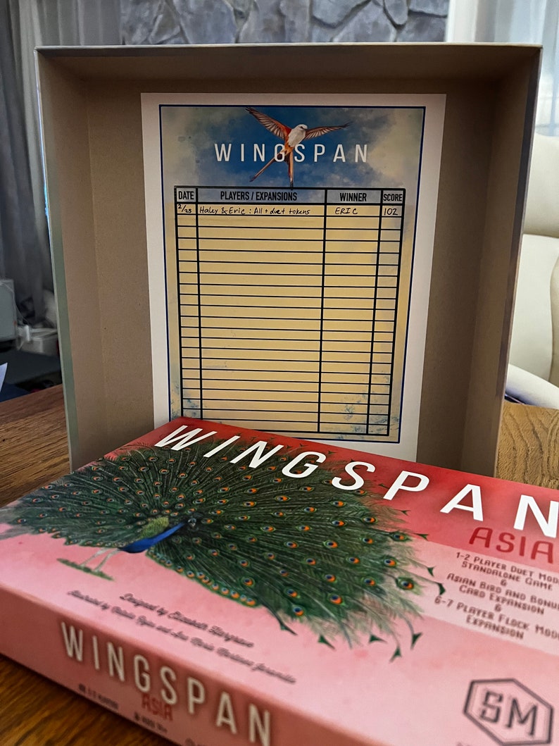 Puede incluir: Una caja de juego de mesa Wingspan con un fondo rosa y una ilustraci&oacute;n de un pavo real. La caja tiene el texto "Wingspan Asia" y un logotipo de Stonemaier Games. La caja est&aacute; abierta y se puede ver una hoja de puntuaci&oacute;n en el interior.