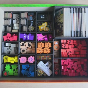 May include: A board game box organiser containing various game components including stacks of white cards, small wooden tokens in red, pink, orange, yellow, green, blue, and purple, and a small white card with the text KONIEC TAHU printed on it.