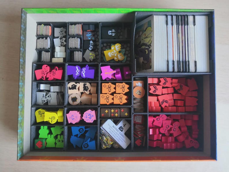 May include: A board game box organiser containing various game components including stacks of white cards, small wooden tokens in red, pink, orange, yellow, green, blue, and purple, and a small white card with the text KONIEC TAHU printed on it.