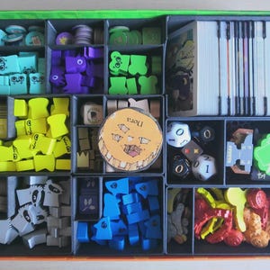 May include: A board game box organiser containing various game components including stacks of white cards, multi-coloured wooden tokens in shapes like ghosts and trees, several polyhedral dice, and a circular cardboard token with the text Nora.