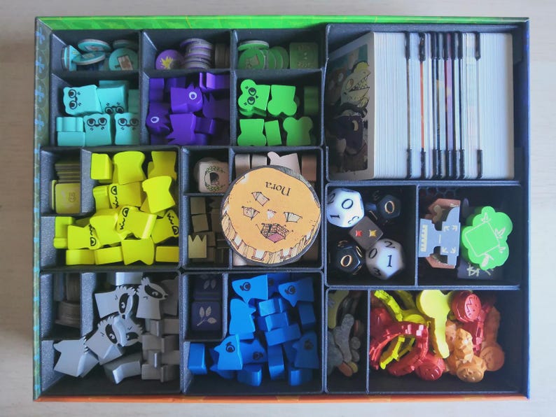 May include: A board game box organiser containing various game components including stacks of white cards, multi-coloured wooden tokens in shapes like ghosts and trees, several polyhedral dice, and a circular cardboard token with the text Nora.