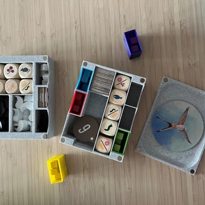 Puede incluir: Un conjunto de cajas grises forradas de fieltro para organizar las piezas de juego. Las cajas contienen varios dados de madera, fichas y cartas. Una caja tiene un inserto redondo con una ilustraci&oacute;n de un p&aacute;jaro.