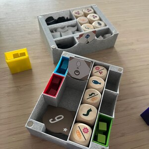 Puede incluir: Una caja de almacenamiento gris con compartimentos para organizar las piezas del juego. La caja contiene dados de madera con varios s&iacute;mbolos, incluyendo un pez, un caracol, una flor y una espiga de trigo. Tambi&eacute;n hay peque&ntilde;as piezas de pl&aacute;stico y una tarjeta con el n&uacute;mero "9" en ella.