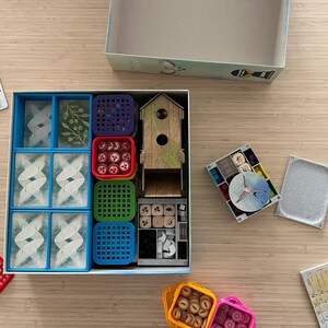 Puede incluir: Una caja de juego azul y blanca con una variedad de compartimentos coloridos para guardar las piezas del juego. Los compartimentos contienen peque&ntilde;os dados de madera, fichas rojas y blancas, y bandejas de pl&aacute;stico azules y verdes. La caja tambi&eacute;n incluye una casita de p&aacute;jaros de madera y un peque&ntilde;o tablero de juego redondo blanco y azul.