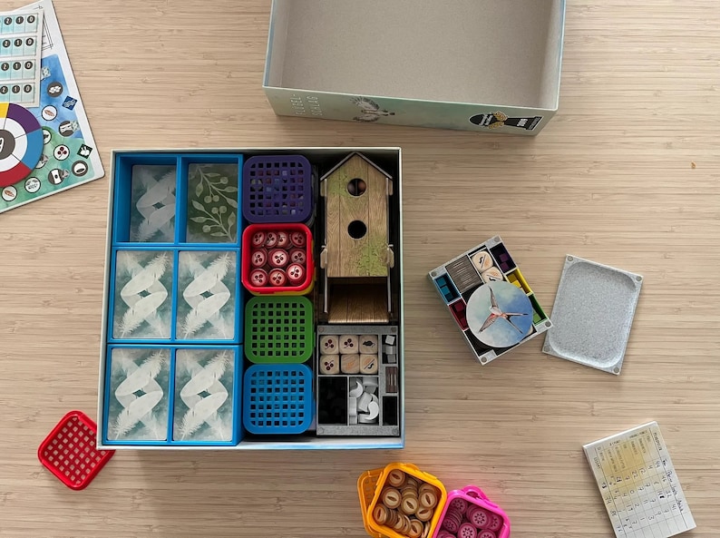 Puede incluir: Una caja de juego azul y blanca con una variedad de compartimentos coloridos para guardar las piezas del juego. Los compartimentos contienen peque&ntilde;os dados de madera, fichas rojas y blancas, y bandejas de pl&aacute;stico azules y verdes. La caja tambi&eacute;n incluye una casita de p&aacute;jaros de madera y un peque&ntilde;o tablero de juego redondo blanco y azul.