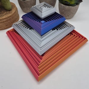 May include: A multi-layered, square-shaped nesting object featuring a gradient of colors from pink and orange at the base to blue and light gray at the top. The structure consists of several concentric, open-frame square tiers stacked vertically.