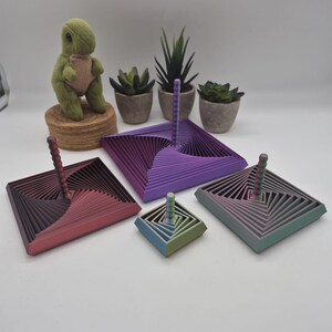May include: Four square-shaped spinning tops with a tiered, geometric, spiral-like design. Each top features a central vertical handle and a multi-colored gradient finish, ranging from red-purple to green-blue, displayed in varying sizes.