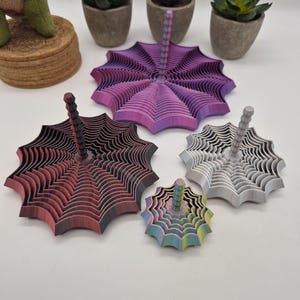 May include: Four 3D printed spinning tops with a spider web design. One top features a purple and pink gradient, one is black and red, one is silver, and the smallest is a multi-colored rainbow gradient. Each top has a central vertical handle.