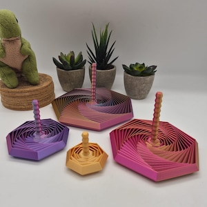 May include: Four hexagonal spinning tops with a geometric, layered, spiral design and a central vertical handle. The tops feature a gradient colour scheme ranging from purple and blue to orange, yellow, and pink, arranged in varying sizes on a surface.