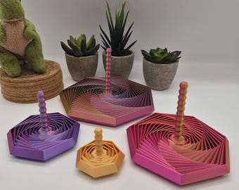 May include: Four hexagonal spinning tops with a geometric, layered, spiral design and a central vertical handle. The tops feature a gradient color scheme ranging from purple and blue to orange, yellow, and pink, arranged in varying sizes on a surface.