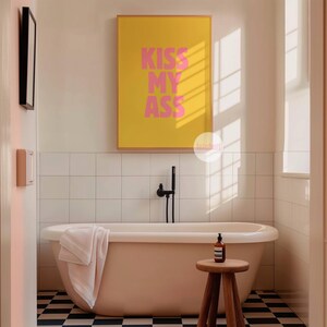 May include: A bathroom scene featuring a freestanding bathtub, a wooden stool, and a framed poster with the text "KISS MY ASS" in pink letters on a yellow background. The floor has a black and white checkered pattern.