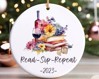 May include: White ceramic Christmas ornament with a watercolor design of a wine bottle, a glass of wine, books, and flowers. The text "Read-Sip-Repeat -2023-" is printed on the ornament. A gold string is attached for hanging.