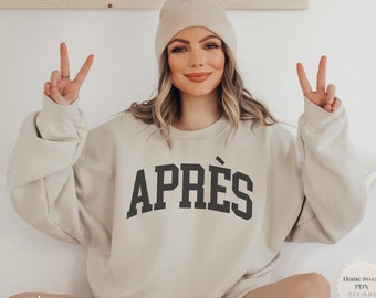 May include: A beige sweatshirt with the word "APRÈS" printed in black on the front. The sweatshirt is being modeled by a person wearing a beige beanie.