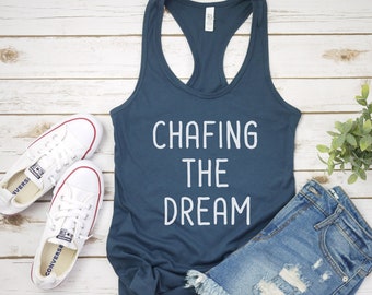 May include: A navy blue tank top with the text "Chafing the Dream" in white lettering. The tank top is laying on a white wooden surface with a pair of white sneakers and a pair of blue denim shorts.