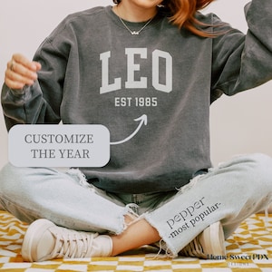 Puede incluir: Una sudadera gris con la palabra "LEO" en letras blancas y el texto "EST 1985" debajo. La sudadera la lleva puesta una persona sentada. El texto "CUSTOMIZE THE YEAR" es visible en un cuadro blanco.