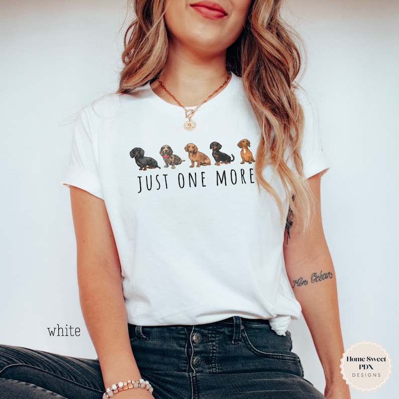 Puede incluir: Camiseta blanca con una l&iacute;nea de perros salchicha y el texto "Just One More".