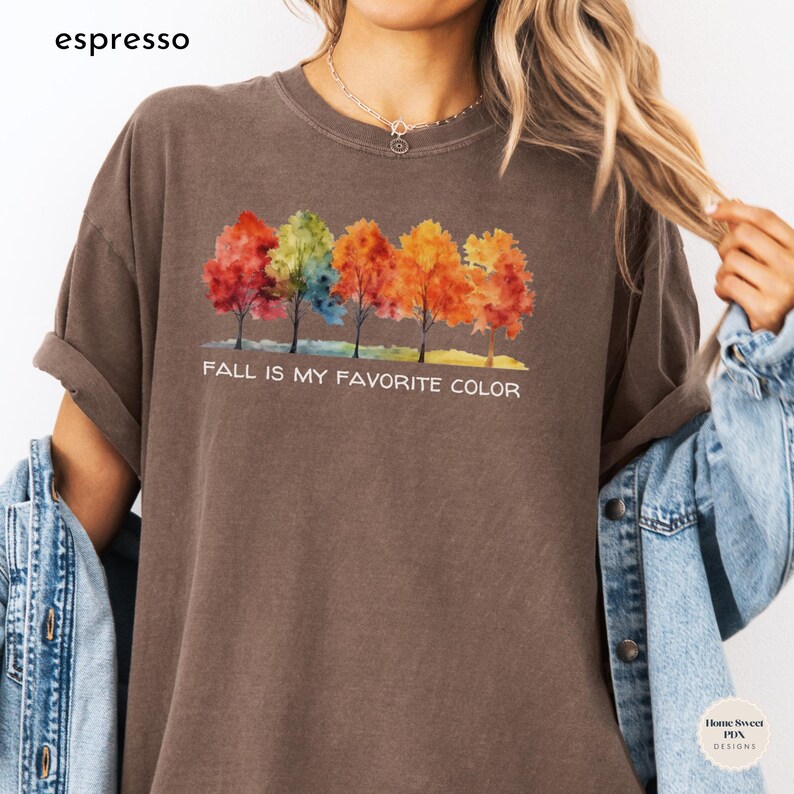 May include: A brown t-shirt with a watercolor design of five colorful trees and the text "Fall is my favorite color".