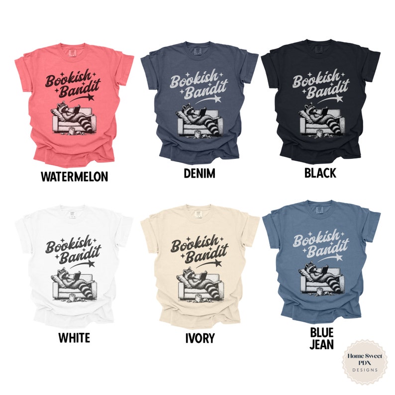 May include: Six different color t-shirts with a graphic of a raccoon reading a book on a couch. The text on the shirts says "Bookish Bandit". The colors of the shirts are watermelon, denim, black, white, ivory, and blue jean.