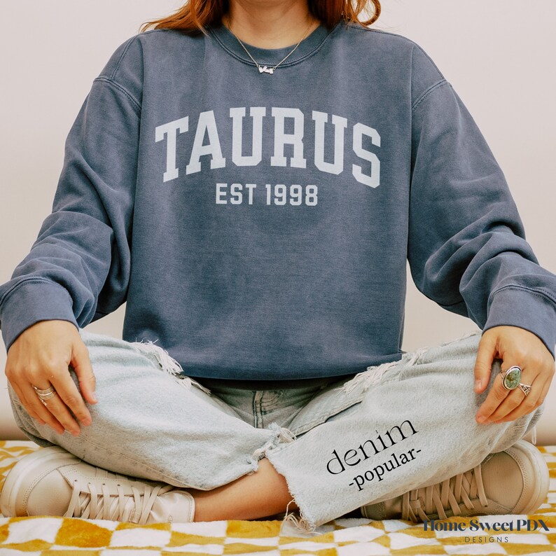 May include: A navy blue sweatshirt with the text "TAURUS EST 1998" in white. The sweatshirt is being worn by a person sitting with their legs crossed.