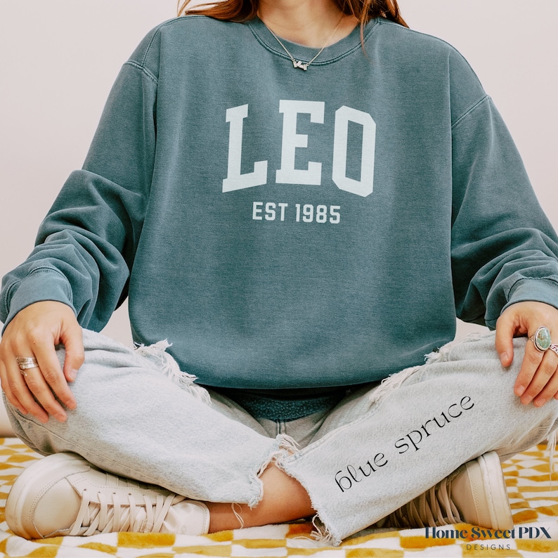 Puede incluir: Una sudadera azul verdosa con el texto "LEO EST 1985" en blanco. La sudadera la lleva puesta una persona sentada con las piernas cruzadas.
