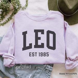 Puede incluir: Una sudadera de color lavanda con la palabra "LEO" en letras negras de bloque y el texto "EST 1985" debajo.