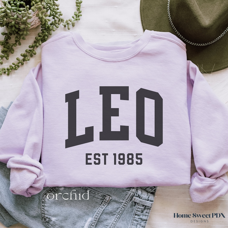 Puede incluir: Una sudadera de color lavanda con la palabra "LEO" en letras negras de bloque y el texto "EST 1985" debajo.