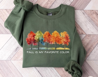 May include: A military green crewneck sweatshirt featuring a watercolor graphic of five trees with autumn-colored foliage in shades of red, orange, and yellow. Below the trees, the text reads, "FALL IS MY FAVORITE COLOR" in white capital letters.